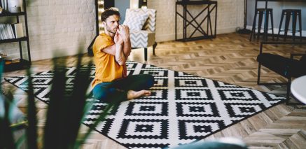 Young concentrated male in orange t shirt and blue pants doing lotus pose with twisting arms and crossed legs on carpet in room